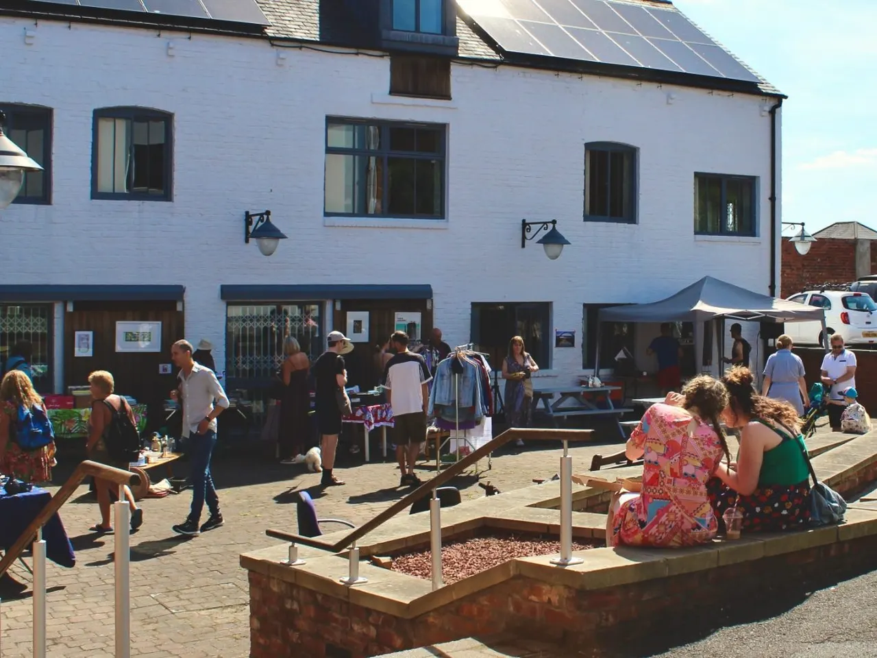 Visitors gather in a sunny courtyard outside a white-painted stone building with solar panels, browsing stalls and mingling at an outdoor event.