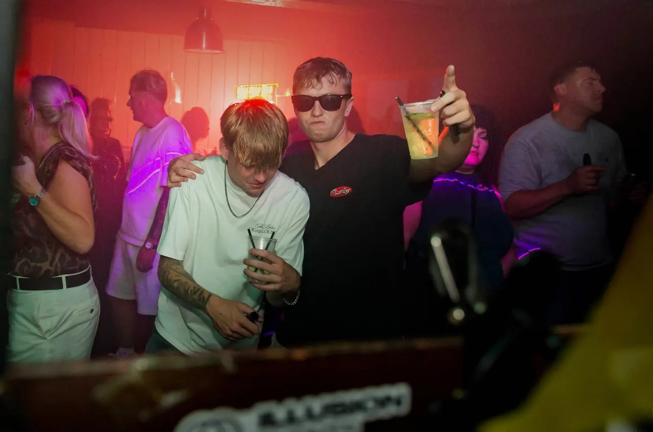 Group of people dancing and socialising in a nightclub lit with red and purple neon lighting.