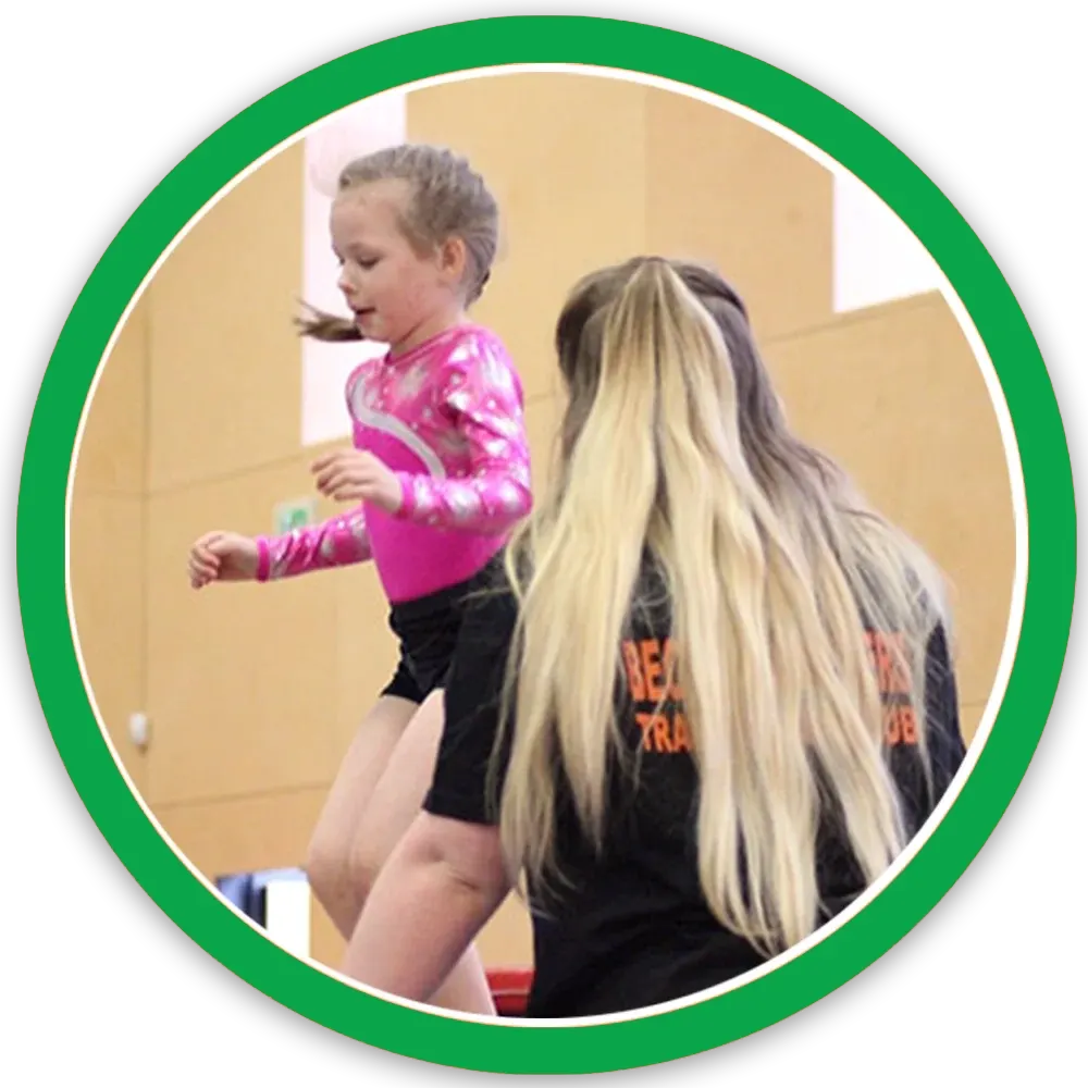 An instructor with long blonde hair watches a young child in a pink top bouncing on a trampoline indoors.