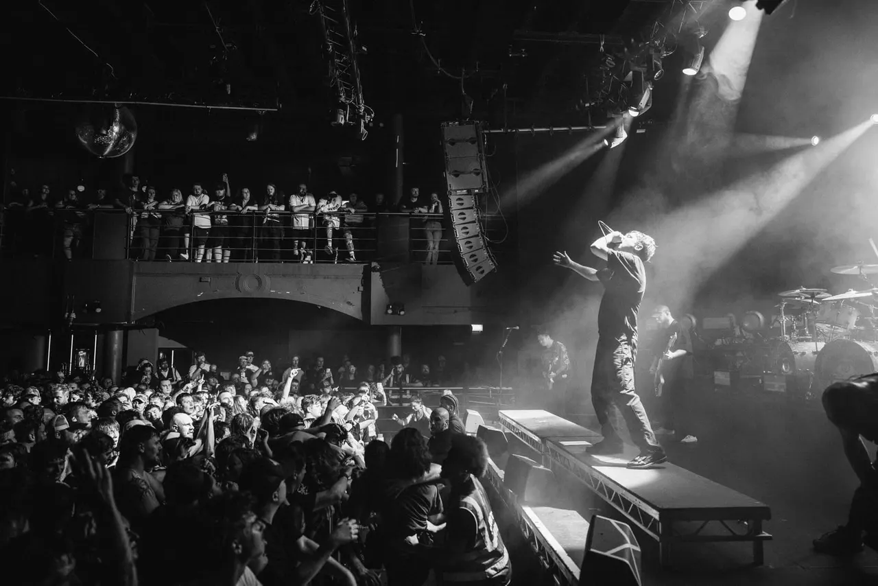 Performer on stage singing to a large crowd in an indoor arena with dramatic lighting and multiple levels of spectators.
