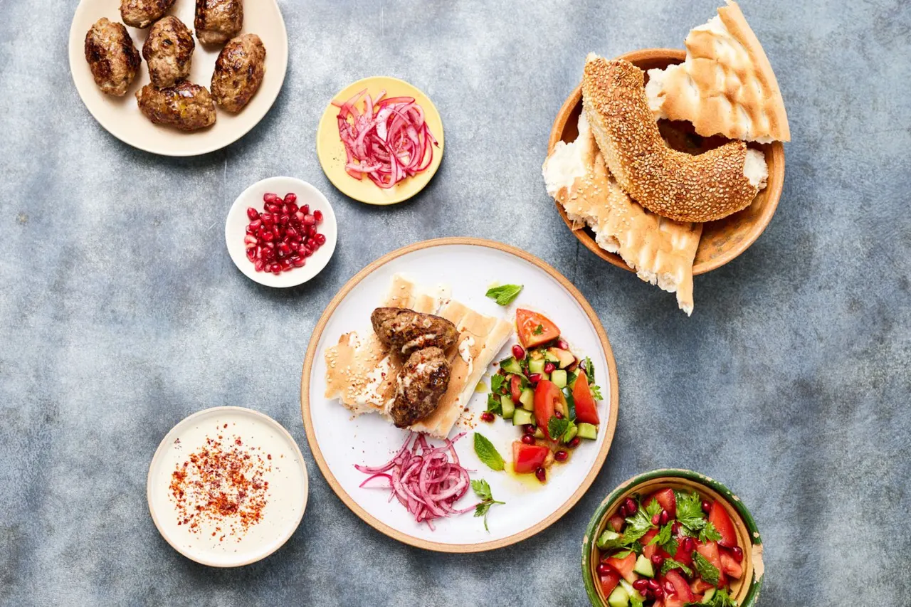 Cooked meat in a seeded bagel with pickled onions, fresh salad and accompaniments arranged on a grey surface.