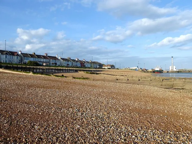 Shingle beach with residential properties and industrial structures visible along the seafront under a partly cloudy sky.