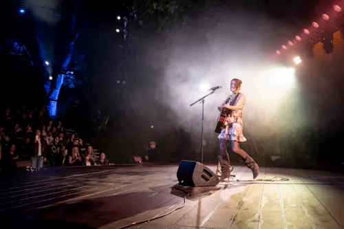 Performer singing at a microphone on a brightly lit stage with an audience seated in darkness below.
