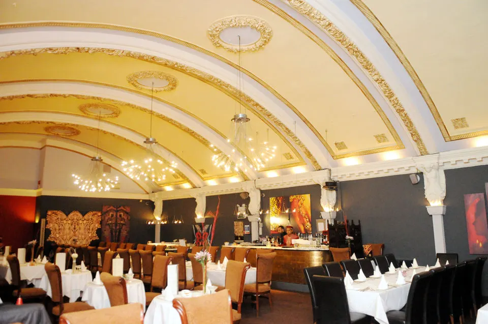 Ornate dining room with gold and white coffered ceiling, set tables with white linens, and decorative artwork on dark walls.