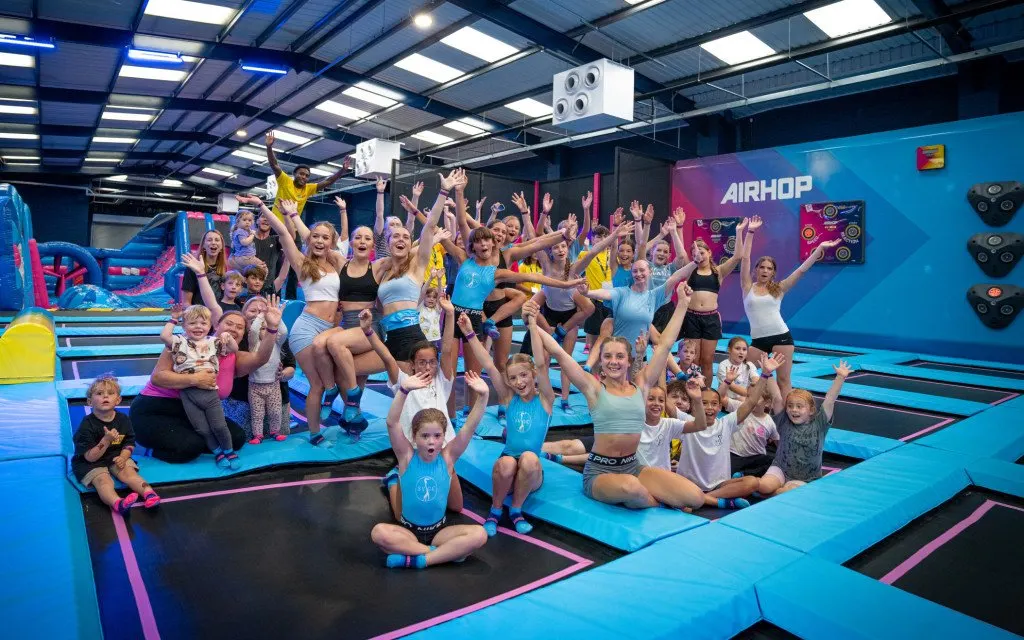 Large group of children and adults posing happily on colourful interconnected trampolines in an indoor trampoline park.