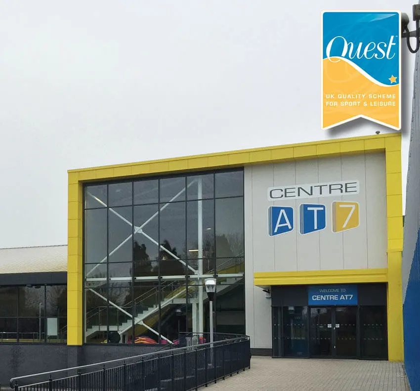Exterior of a modern sports leisure centre with yellow and white panelling, large windows, and a Quest quality scheme badge.