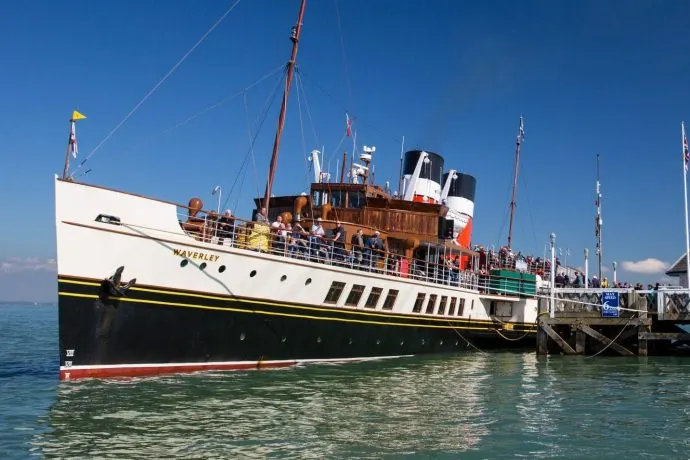 Paddle steamer moored at a dock with passengers aboard under clear blue sky.