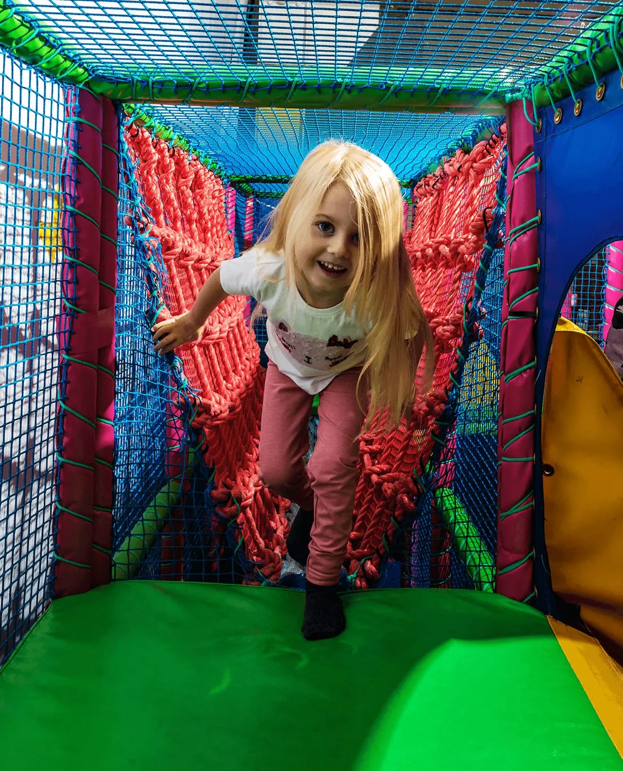 Young girl crawling through a colourful indoor soft play tunnel with red netting and foam obstacles.