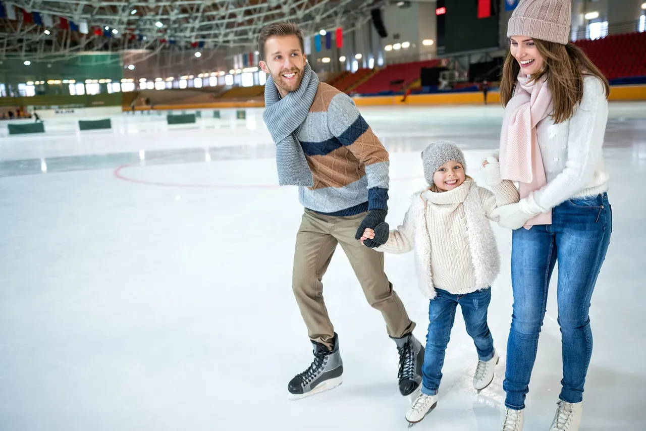 Family of four ice skating together at an indoor ice rink, smiling at the camera.