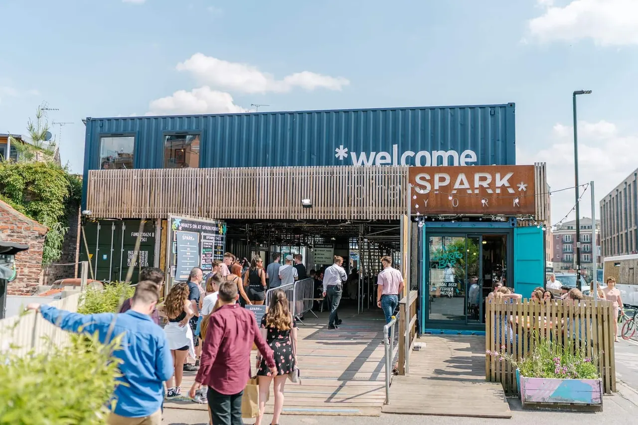 Visitors arrive at a modern waterfront venue with shipping container architecture, stepping onto a wooden deck with outdoor seating and plants.