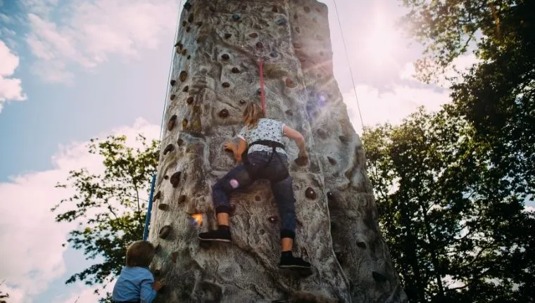 A person rock climbing an outdoor climbing wall with a rope, watched by another visitor below.