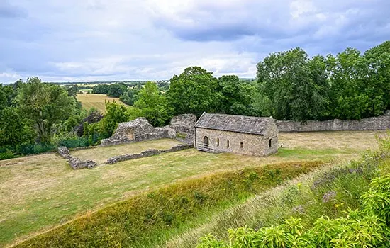 Ruins of a stone fortification on grassy terrain surrounded by trees and rolling countryside under cloudy sky.