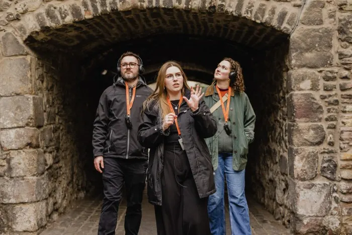 Three people wearing orange lanyards walk through a stone archway during a guided tour.