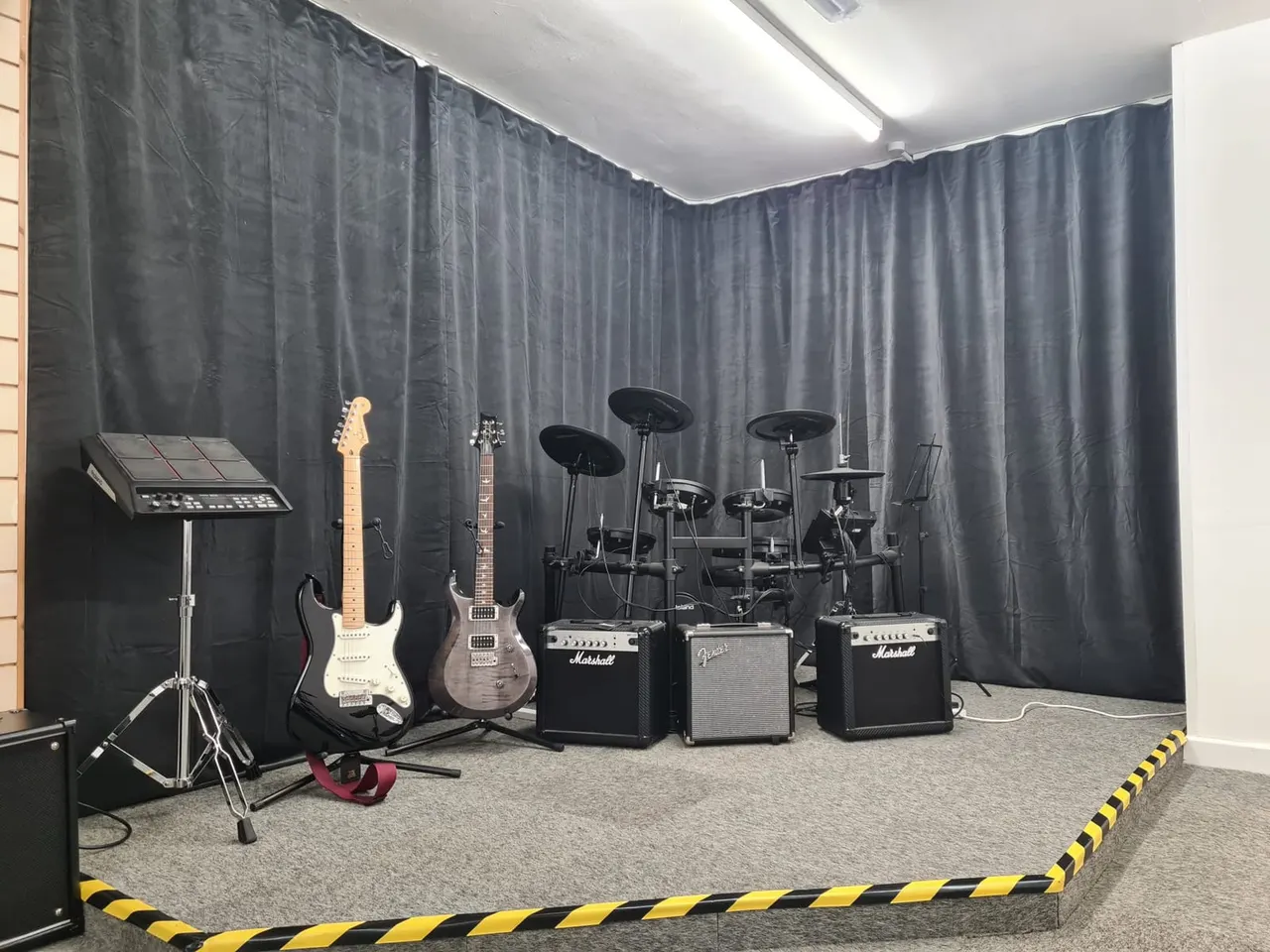 Music rehearsal studio with electric guitars, amplifiers, electronic drums, and keyboard on a stage area with black curtain backdrop.