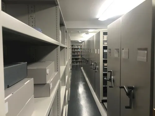 A long, narrow storage or archive corridor lined with white shelving units and filing systems on both sides.