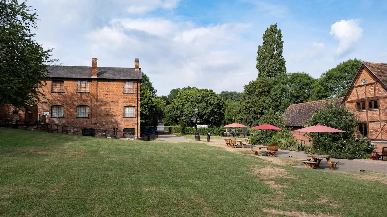 Historic brick buildings with picnic tables and red umbrellas on a grassy courtyard surrounded by trees.