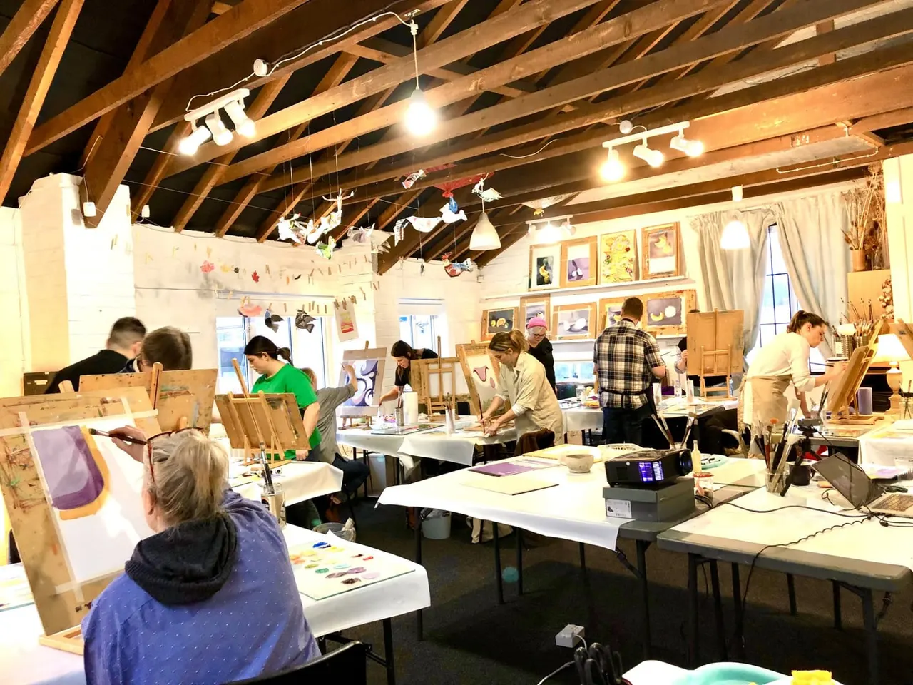 A group of people painting and drawing at easels and tables in a spacious studio with exposed wooden beams and hanging decorations.