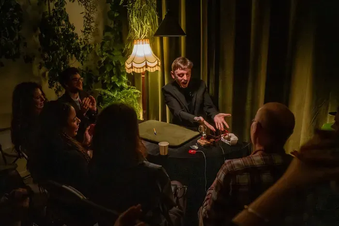 Group of people gathered around a table in a dimly lit room with hanging plants and a decorative lamp, appearing to watch a close-up magic performance.
