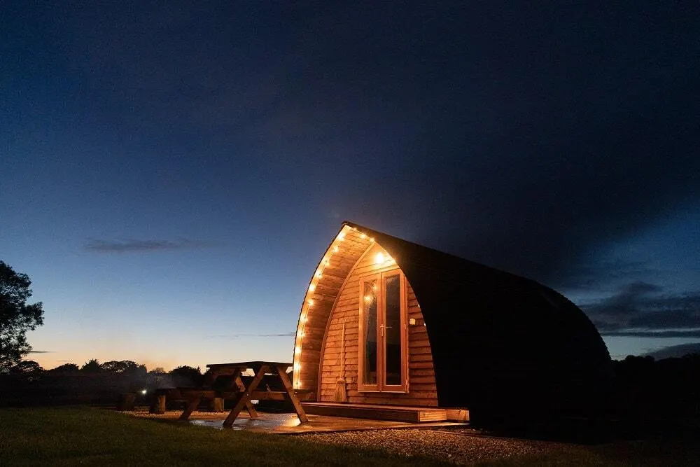 Illuminated wooden glamping pod with arched roof on rural farmland at dusk, with picnic table beside it.