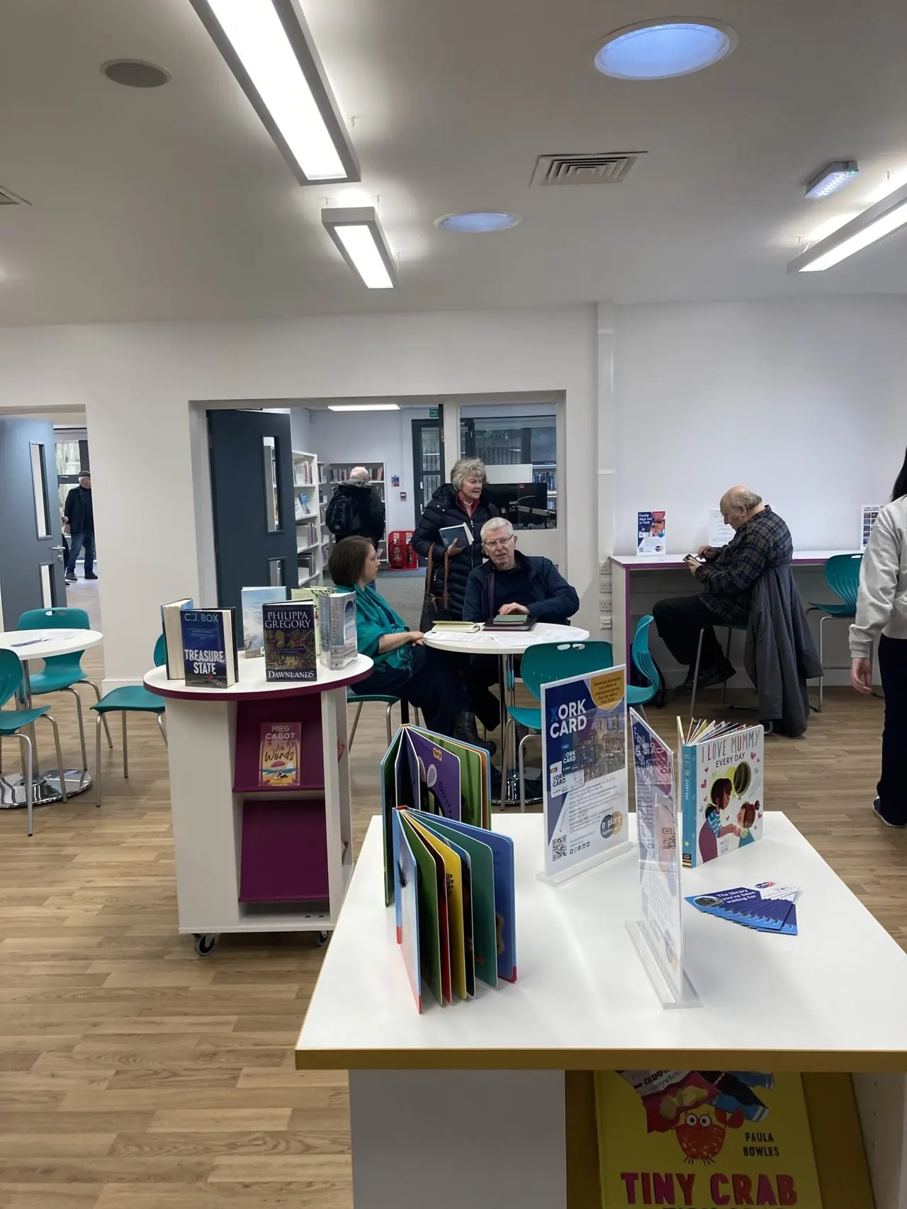 Interior of a modern library with visitors browsing books at tables and displays, staff at a desk, and shelving visible in background.