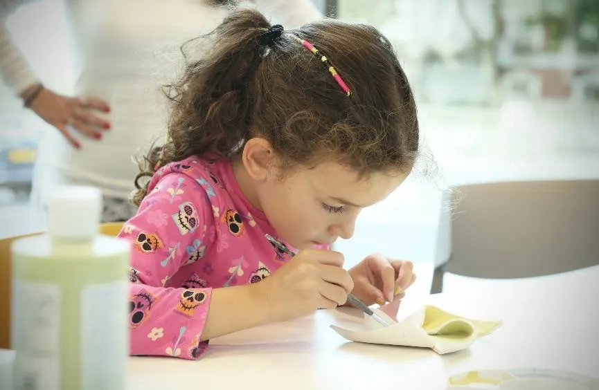 Young child in pink top concentrating while painting or drawing at a table indoors.