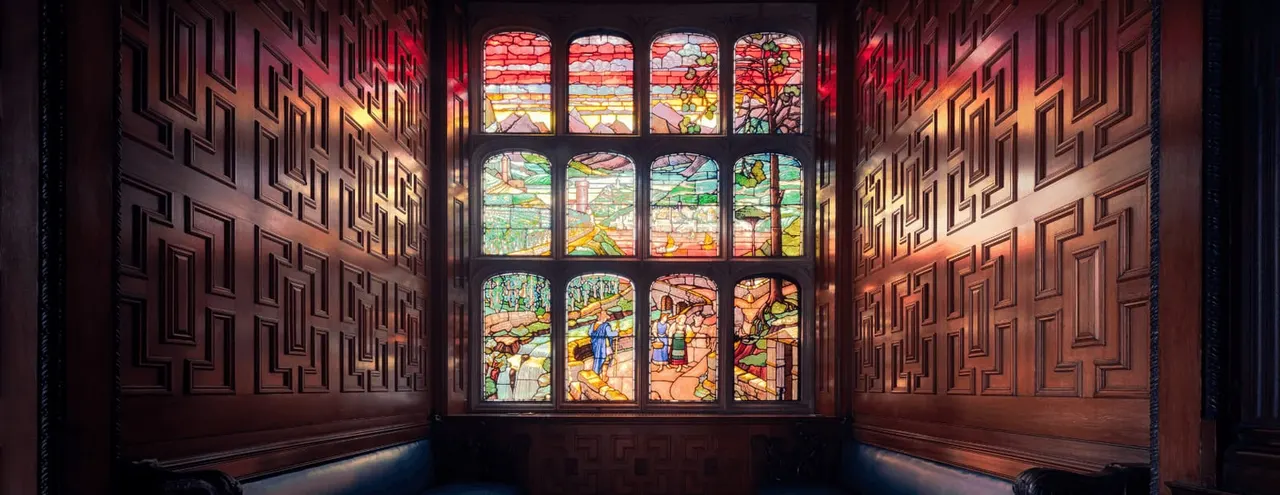 Large stained glass window depicting agricultural and rural scenes, viewed through ornate wooden doorways in a historic interior space.