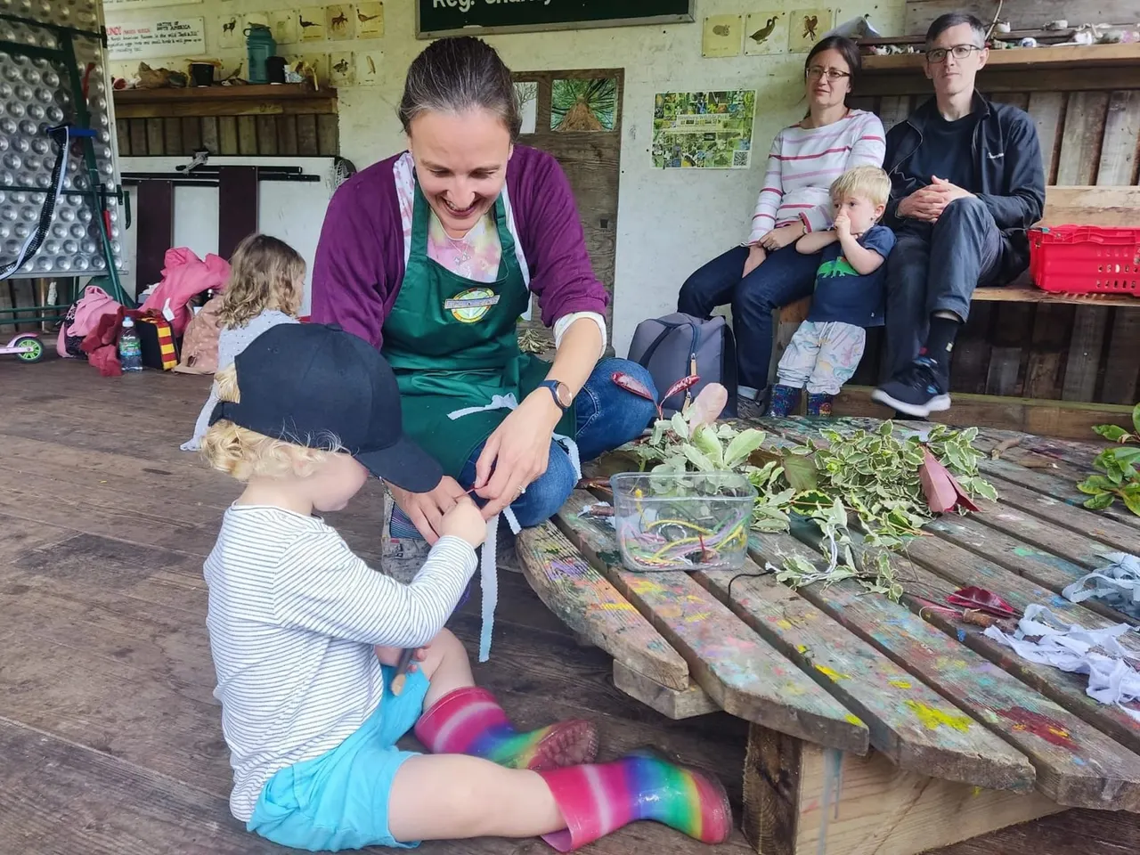 A staff member in a green apron guides young children in a hands-on wildlife educational activity indoors, with families watching from benches nearby.