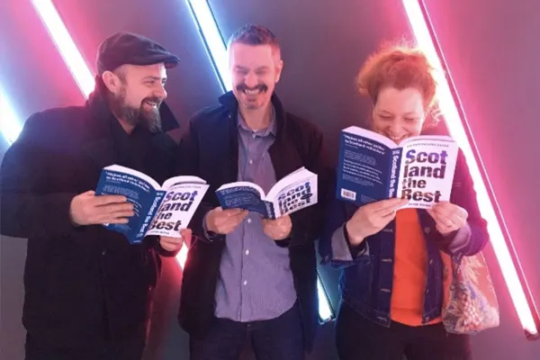 Three people holding books and smiling in front of pink and blue neon lights.