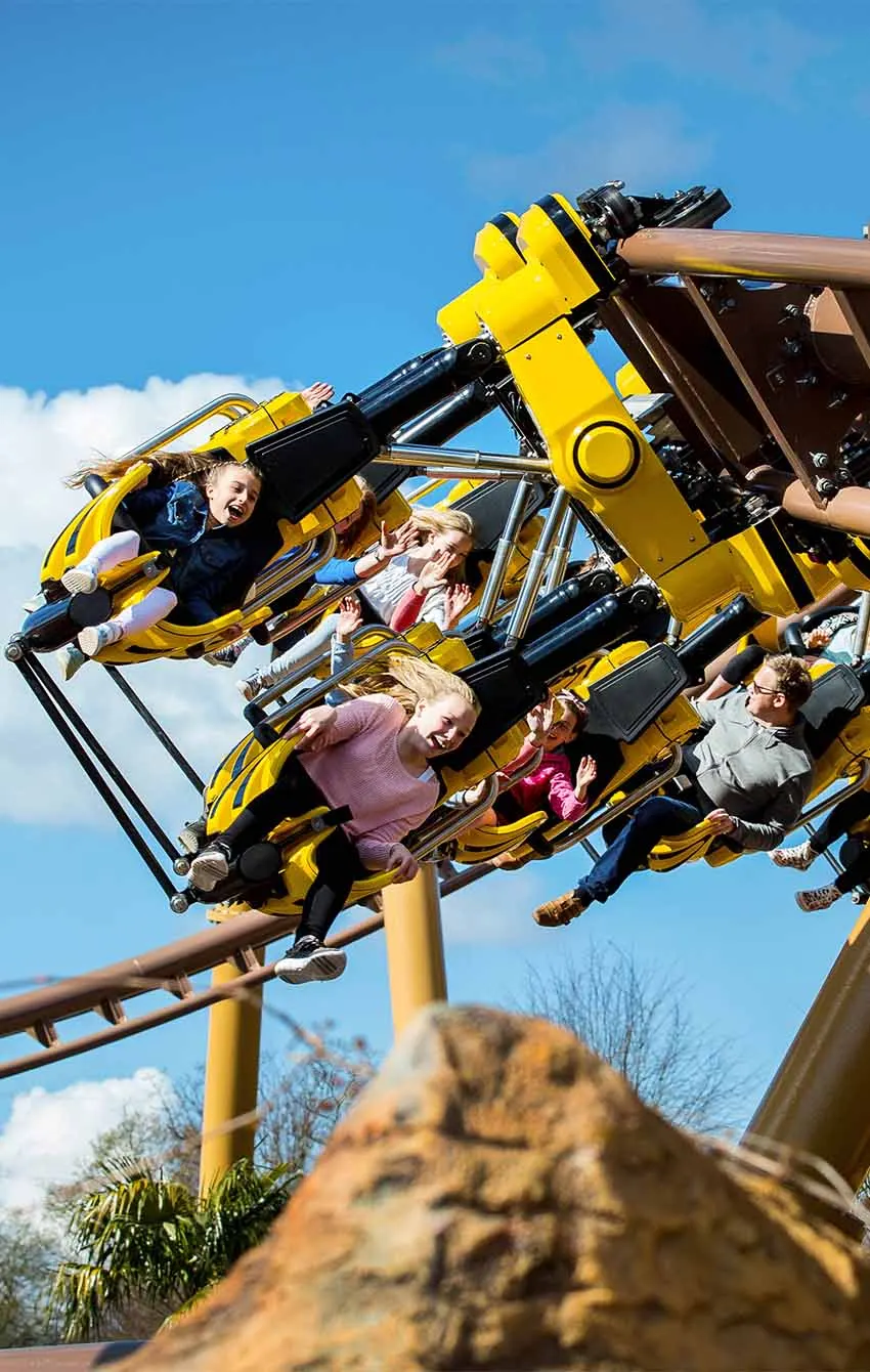 Families and children riding a yellow and black roller coaster with arms raised in excitement against a blue sky.