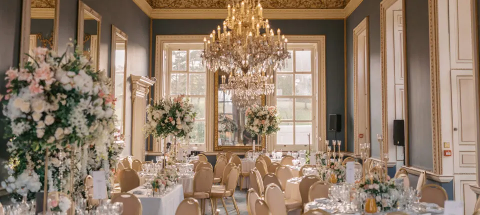 Elegant banquet room with cream and gold chairs, white floral arrangements, crystal chandelier, and formal table settings.