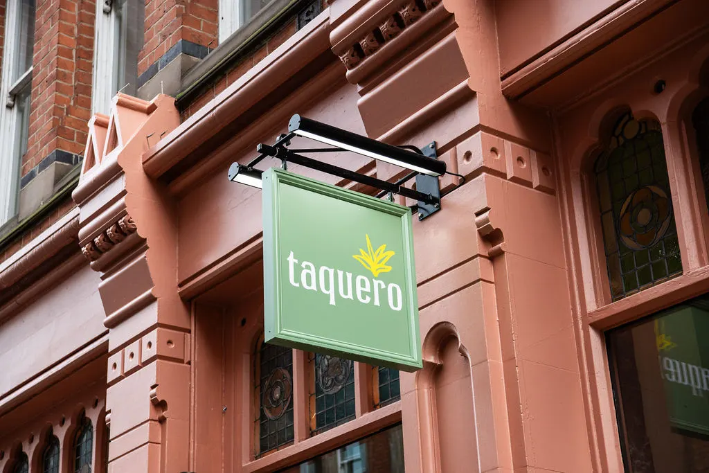 Green illuminated signage for "Taquero" restaurant mounted on a terracotta-coloured Victorian building facade.
