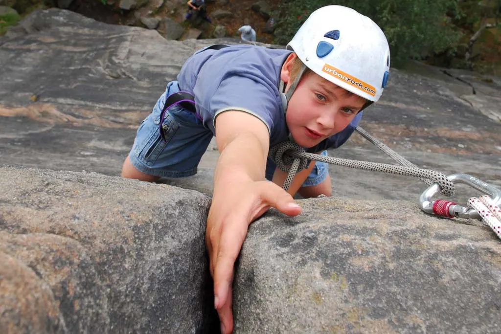 Child in safety helmet and harness rock climbing on an outdoor stone face with rope.