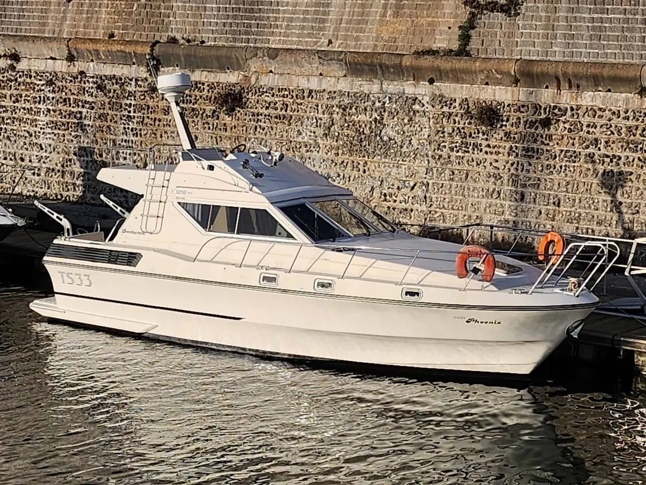 White motor yacht moored at a stone harbour wall with orange lifebuoys visible on deck.