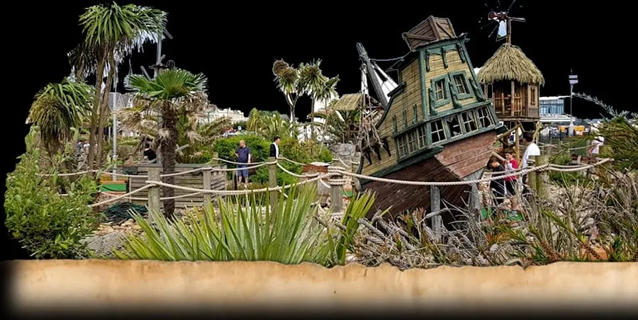 Themed adventure golf course with a shipwrecked pirate ship structure, tropical plants, and visitors playing mini golf.