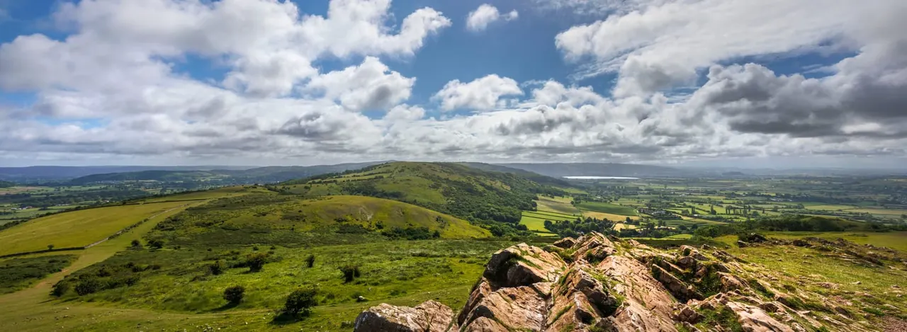 Rolling green hills and distant farmland viewed from a rocky summit under a bright cloudy sky.