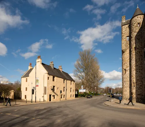 Stone historic building on a street with a church tower, people walking, and bare trees under a blue sky.