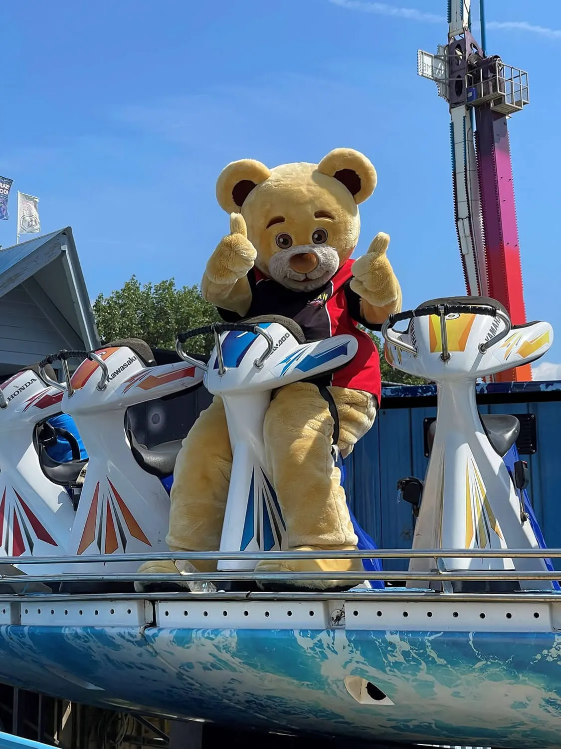 A large teddy bear mascot in a red jacket gives thumbs up while standing on a fairground ride car, with a tall ride tower visible behind.