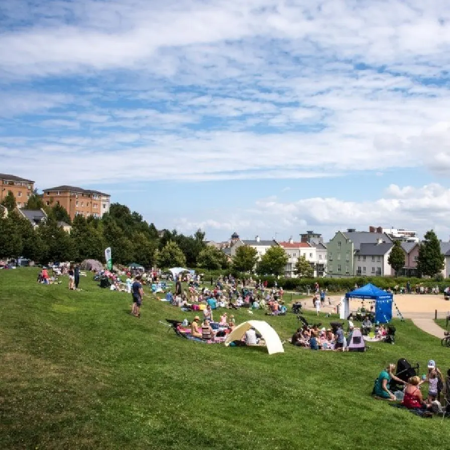 Large grassy park with families and groups scattered across the lawn, buildings and blue event tent visible in the background on a sunny day.
