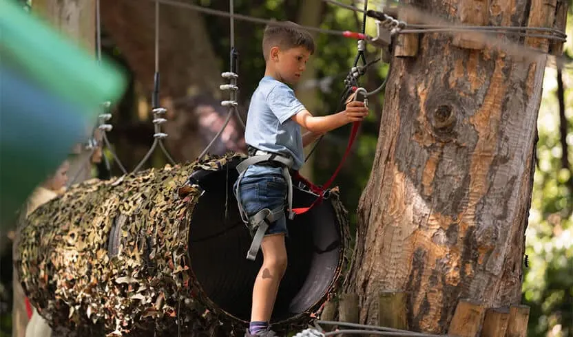 Child climbing through a wooden tunnel course secured with red safety harness and ropes.