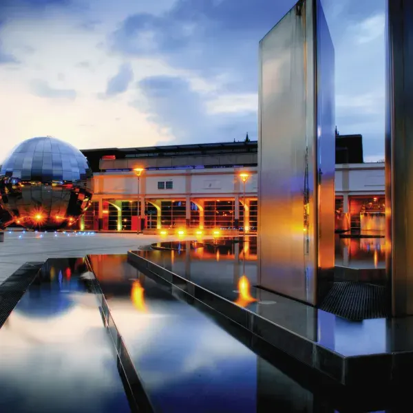 Modern science centre with geodesic dome and illuminated buildings reflected in still water at dusk.