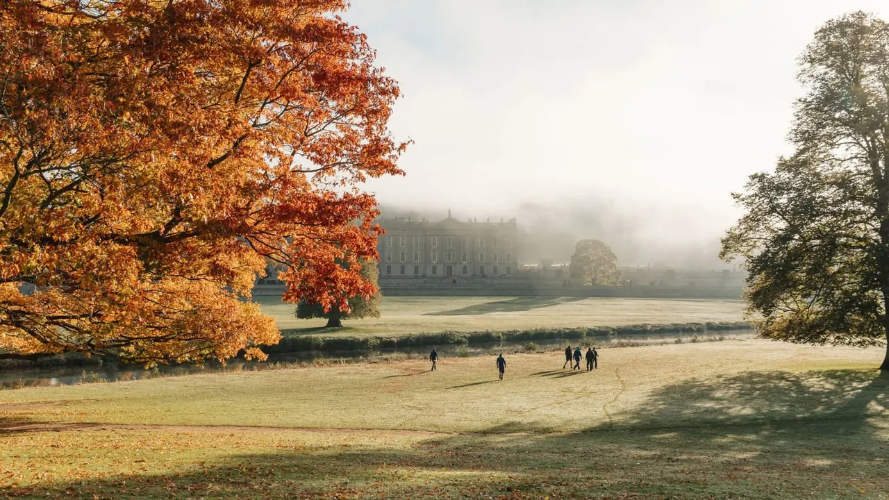 Visitors walking across autumn parkland with a grand mansion visible through mist beyond trees and water.