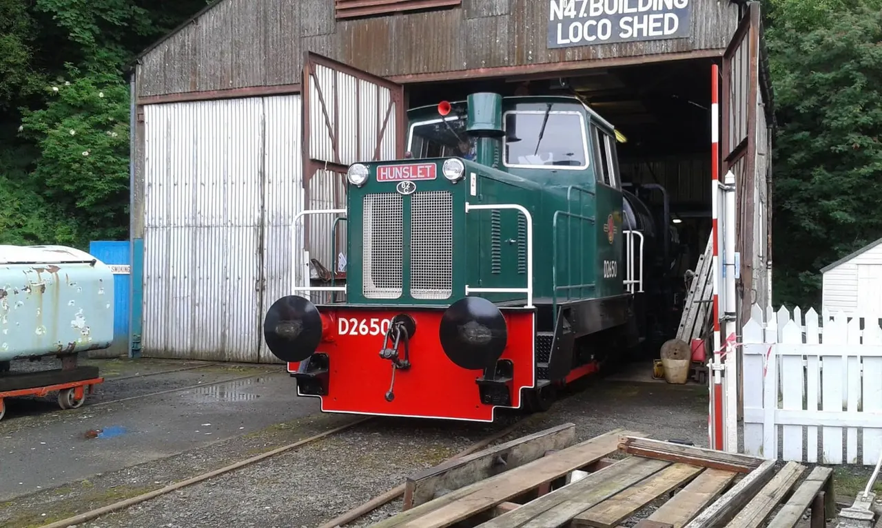 Green and red vintage steam locomotive stored in a metal industrial shed surrounded by railway heritage equipment.