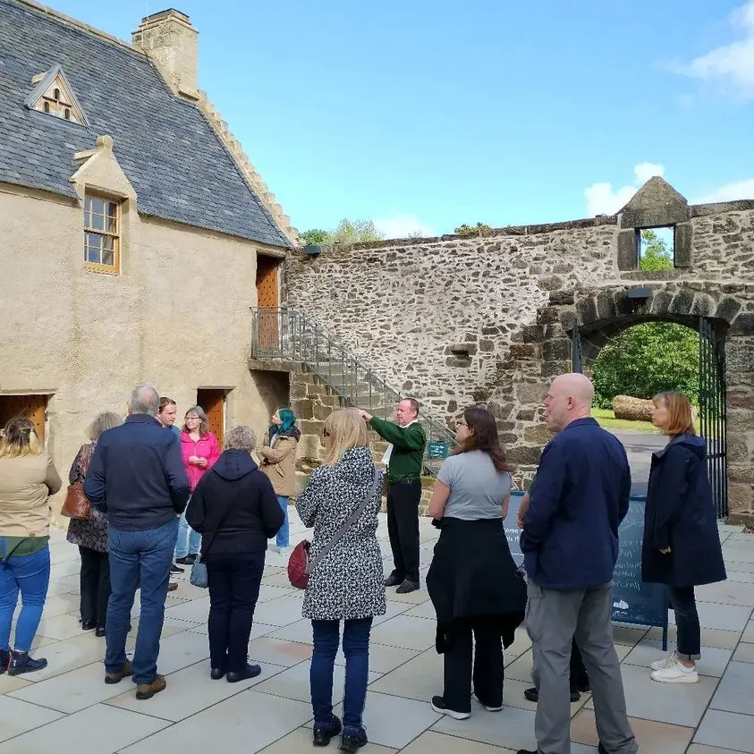 A group of visitors gathered in the courtyard of a historic stone building and ruined walls during a guided tour.