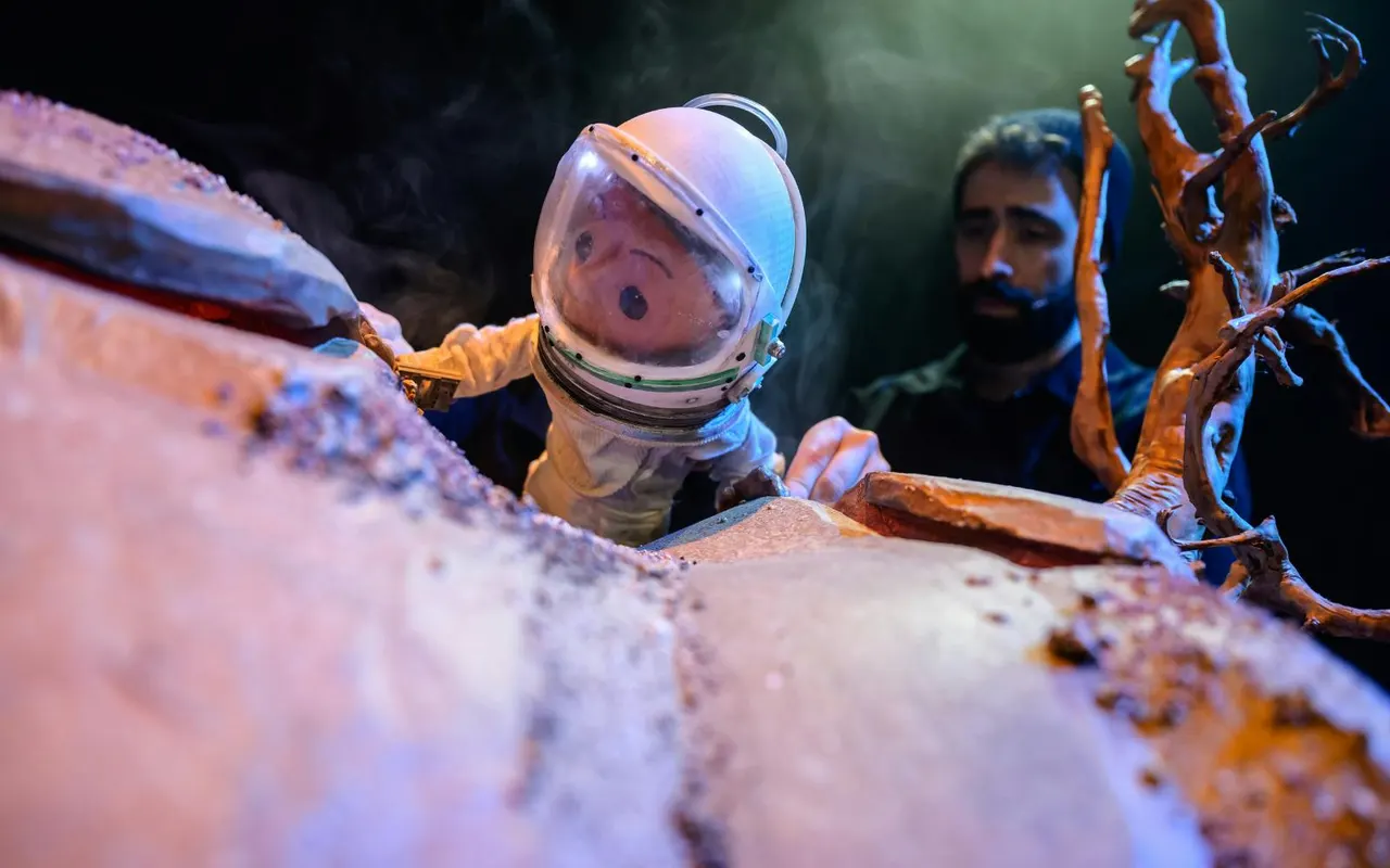 Puppet astronaut in spacesuit exploring rocky terrain with another figure visible in background.