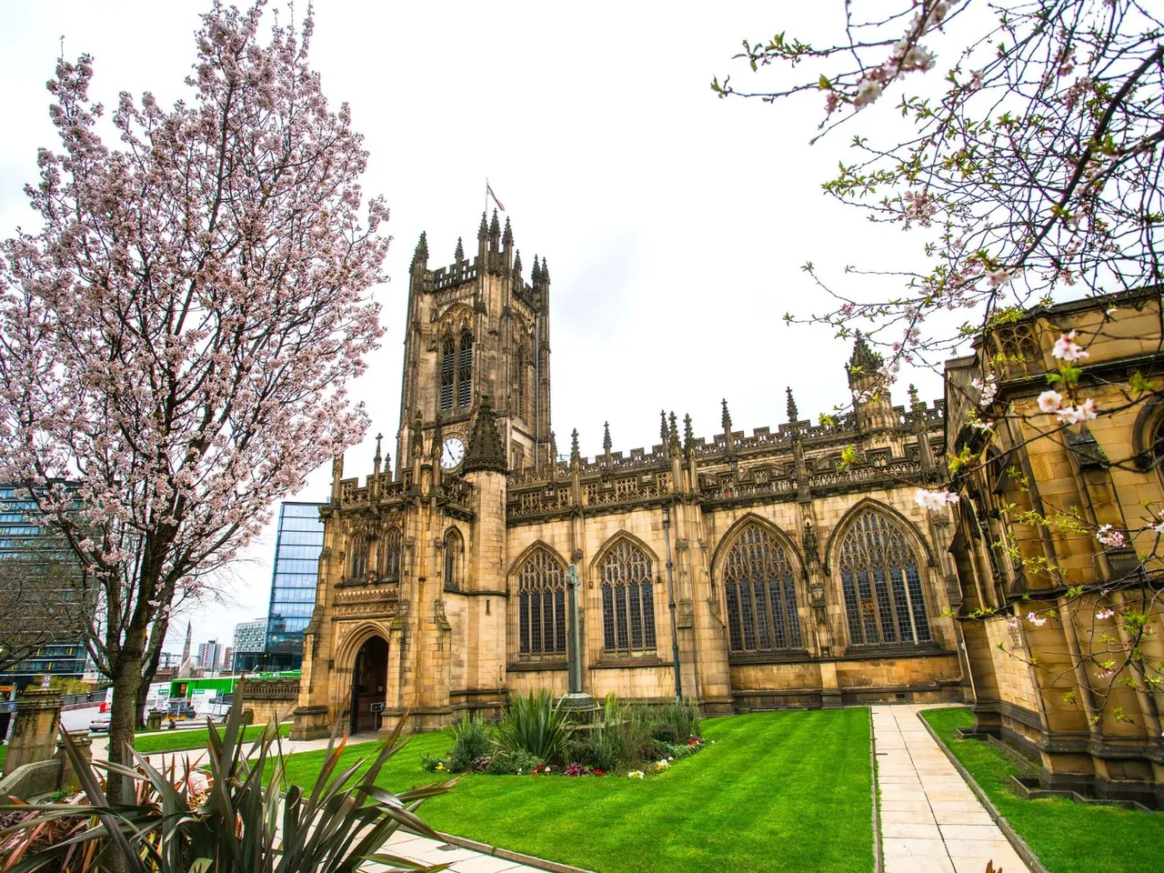 Gothic stone church with tall tower and large windows, surrounded by blooming spring trees and manicured gardens, modern city buildings visible beyond.