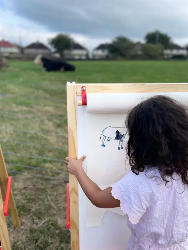 Child drawing a picture of farm animals on an easel in a rural field setting.