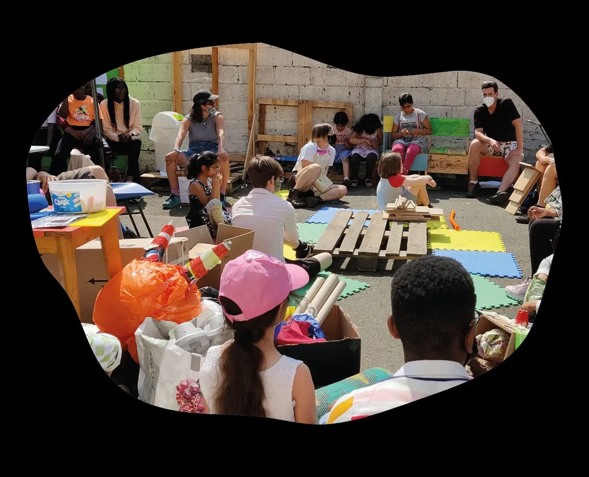 Children and adults participating in a creative workshop with recycled materials and wooden structures in an indoor space.