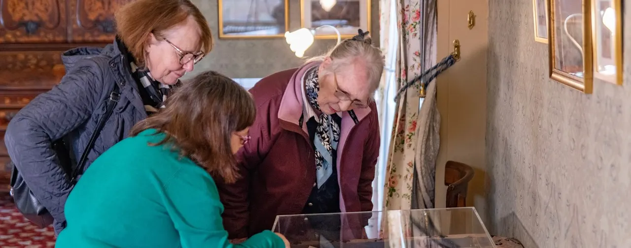 Three visitors examining a display case in a period room with framed pictures and textiles on the walls.