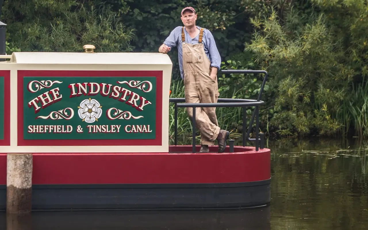 Man in work clothes and cap standing on the deck of a narrowboat named "The Industry" on a canal surrounded by green vegetation.