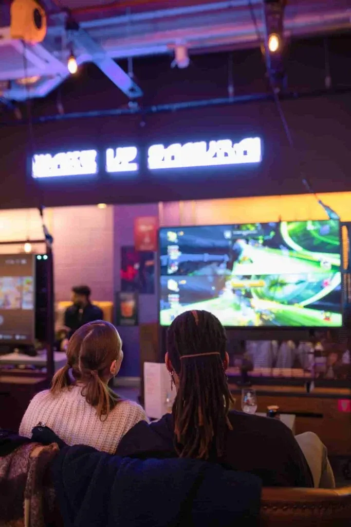 Two people sitting at a counter watching a screen in a dimly lit arcade or gaming venue with neon signage.